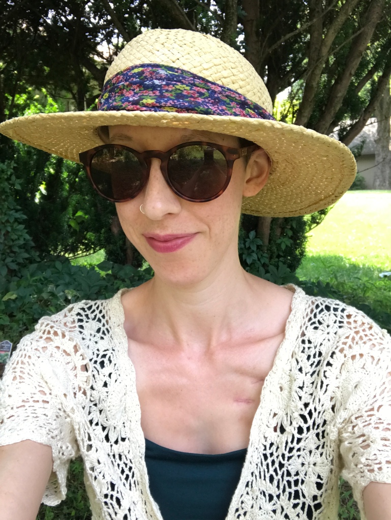 A selfie of Kelly wearing sunglasses, a dark green top, a white crocheted short-sleeved cardigan, and a wide-brimmed straw hat with floral fabric around the brim.