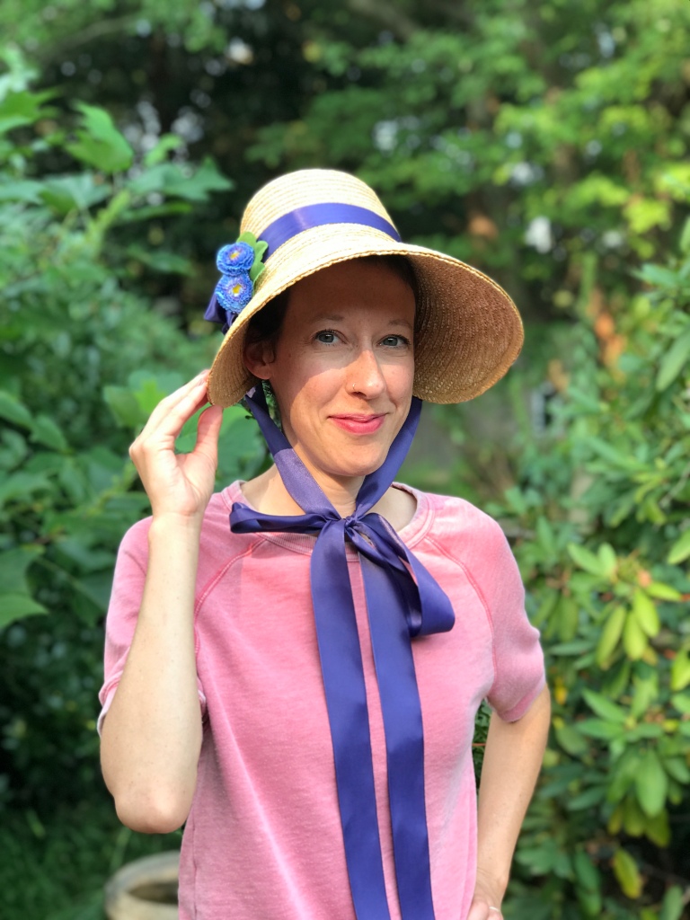 Kelly wears a pink shirt and a straw bonnet with a large brim and a back dome that extends fairly far out. The brim is wrapped with a long purple ribbon (which is tied under Kelly's chin) and blue faux flowers.