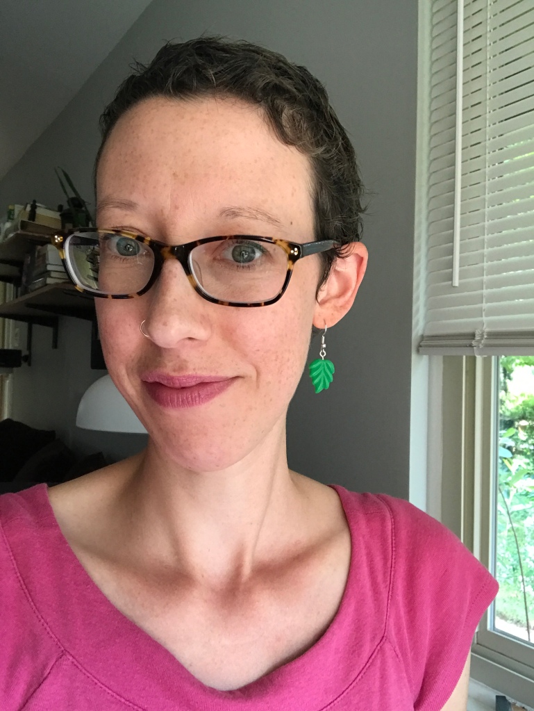 Selfie of Kelly. She's wearing glasses, a pink shirt, and green leaf earrings. Her hair is short but starting to curl.