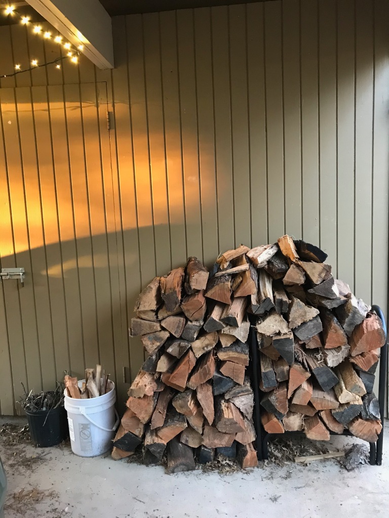 A large stack of firewood leans against a shed. There are a few buckets with kindling in them. The setting sun casts bright orange light over the shed.