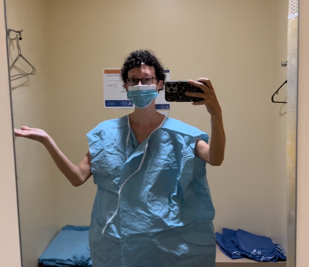 A mirror shot of Kelly (a white woman with short, curly brown hair and glasses) wearing a large blue paper gown.